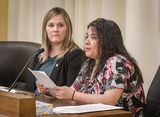 Jacqueline Arias sits at the testifiers' table. Holding a piece of paper in her hands, she speaks to someone out of view. Representative Koegel sits next to her and watches as she speaks.